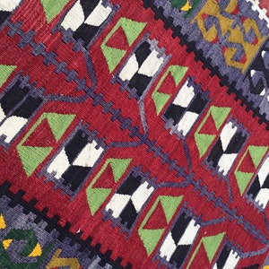 May include: Close-up of a vibrant, handwoven rug with a rich red background. The rug features geometric patterns in black, white, green, and purple, creating a striking visual contrast. The design includes repeating diamond and zigzag motifs.
