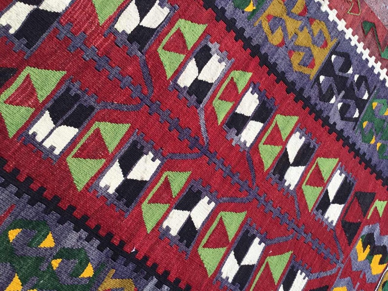 May include: Close-up of a vibrant, handwoven rug with a rich red background. The rug features geometric patterns in black, white, green, and purple, creating a striking visual contrast. The design includes repeating diamond and zigzag motifs.