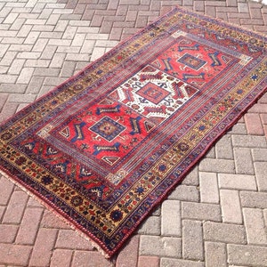 May include: A red, blue, and gold patterned rug with a traditional design. The rug has a fringe border and is laid out on a brick patio.
