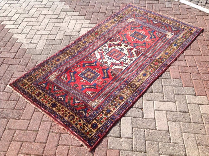May include: A red, blue, and gold patterned rug with a traditional design. The rug has a fringe border and is laid out on a brick patio.