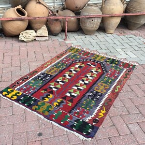 May include: A rectangular, hand-woven rug with a vibrant geometric pattern. The rug features a central design in red, green, and white, framed by borders in yellow, blue, and black. The rug has a fringed edge and is displayed on a brick surface.