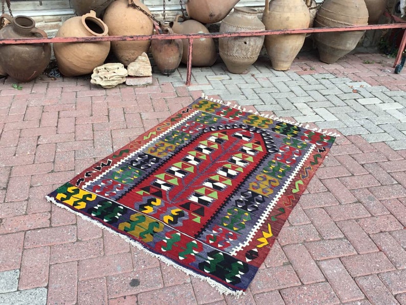 May include: A rectangular, hand-woven rug with a vibrant geometric pattern. The rug features a central design in red, green, and white, framed by borders in yellow, blue, and black. The rug has a fringed edge and is displayed on a brick surface.