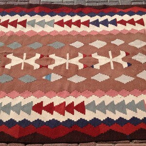 May include: A rectangular, patterned rug with a brown central field. The rug features geometric designs in red, white, blue, and pink. The border has a zig-zag pattern in dark red and blue. The rug is made of woven fabric.