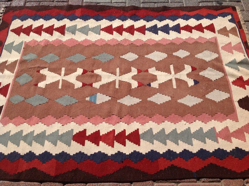 May include: A rectangular, patterned rug with a brown central field. The rug features geometric designs in red, white, blue, and pink. The border has a zig-zag pattern in dark red and blue. The rug is made of woven fabric.