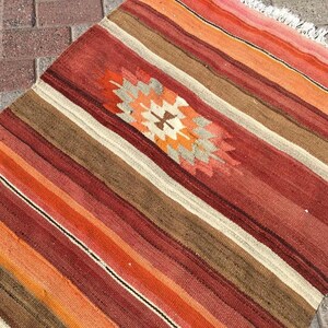 May include: A rectangular, striped rug with a central geometric design. The rug features alternating stripes of red, orange, brown, and cream colors. The geometric design in the center has orange, cream, and gray colors. The rug has a fringed edge.