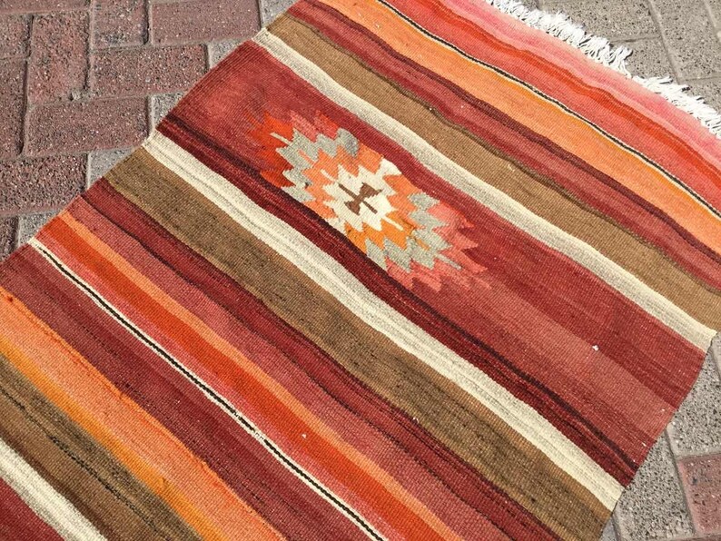 May include: A rectangular, striped rug with a central geometric design. The rug features alternating stripes of red, orange, brown, and cream colors. The geometric design in the center has orange, cream, and gray colors. The rug has a fringed edge.