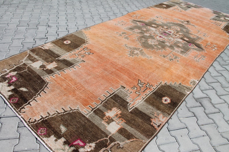 May include: An orange and brown vintage rug with a central medallion design. The rug features geometric patterns in shades of brown, beige, and pink. The rug is rectangular and appears to be made of woven material.