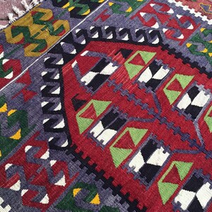 May include: A hand-woven rug with a geometric pattern. The rug features a red central field with black, white, and green geometric shapes. The border is a mix of purple, green, and yellow, with white fringe.