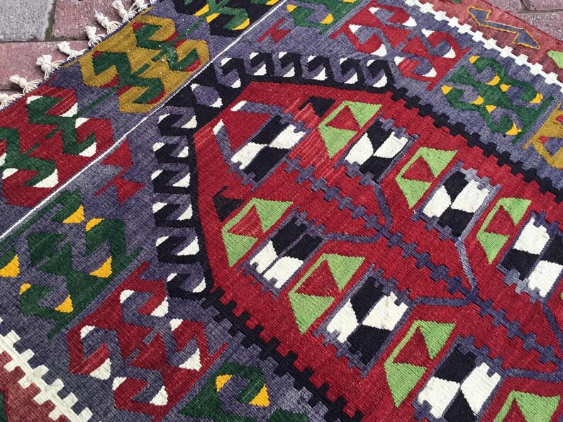 May include: A hand-woven rug with a geometric pattern. The rug features a red central field with black, white, and green geometric shapes. The border is a mix of purple, green, and yellow, with white fringe.