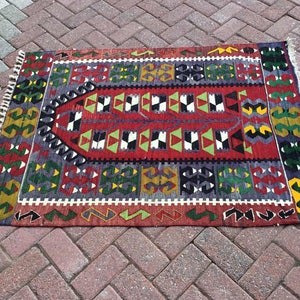 May include: A rectangular, hand-woven rug with a central red field and geometric patterns in green, white, black, and yellow. The rug has a border with repeating shapes and fringed edges. The rug is approximately 36 inches long and 24 inches wide.