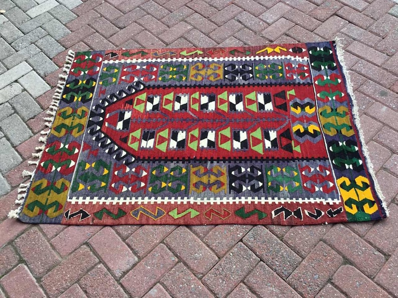 May include: A rectangular, hand-woven rug with a central red field and geometric patterns in green, white, black, and yellow. The rug has a border with repeating shapes and fringed edges. The rug is approximately 36 inches long and 24 inches wide.