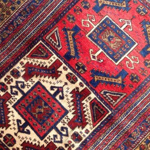 May include: A close-up of a traditional rug with intricate geometric patterns in shades of red, blue, and white. The rug features a central medallion with a repeating design of stylized flowers and leaves.