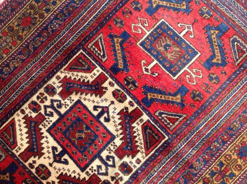 May include: A close-up of a traditional rug with intricate geometric patterns in shades of red, blue, and white. The rug features a central medallion with a repeating design of stylized flowers and leaves.