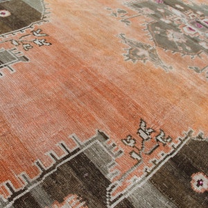 May include: An antique, distressed rug with a warm color palette. The rug features a central field of faded orange, bordered by geometric patterns in shades of brown, gray, and beige. The design includes floral and geometric motifs.