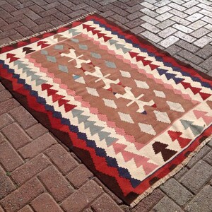 May include: Rectangular woven rug with a geometric pattern. The rug features a brown central field with white, pink, and gray diamond shapes. The border is a mix of red, blue, and brown triangles and lines. The rug has a fringed edge.