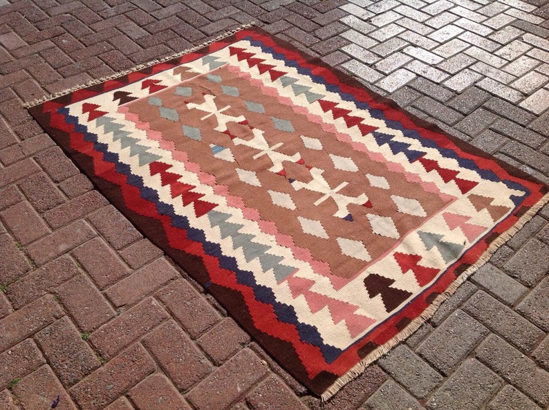 May include: Rectangular woven rug with a geometric pattern. The rug features a brown central field with white, pink, and gray diamond shapes. The border is a mix of red, blue, and brown triangles and lines. The rug has a fringed edge.