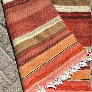 May include: A handwoven rug with a striped pattern in shades of red, orange, brown, and cream. The rug has a fringed edge and is partially rolled up, revealing the underside. The rug is likely made of natural fibers.