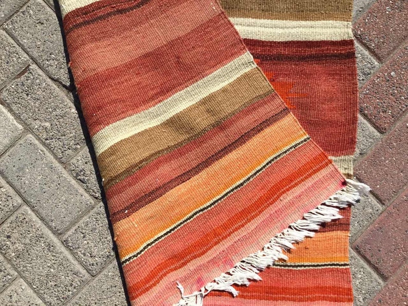 May include: A handwoven rug with a striped pattern in shades of red, orange, brown, and cream. The rug has a fringed edge and is partially rolled up, revealing the underside. The rug is likely made of natural fibers.