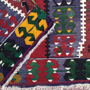 May include: A folded, colorful rug with a geometric pattern. The rug features a combination of red, blue, green, yellow, and white designs. The edges are finished with a white fringe.
