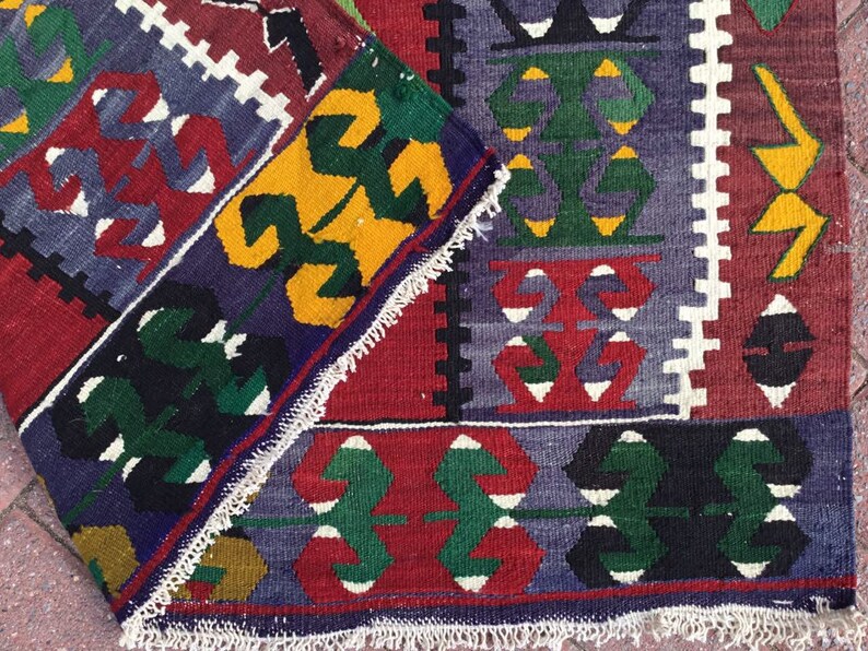 May include: A folded, colorful rug with a geometric pattern. The rug features a combination of red, blue, green, yellow, and white designs. The edges are finished with a white fringe.