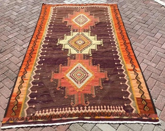 May include: A vintage Turkish rug with a geometric pattern in shades of red, orange, brown, and cream. The rug has a fringed edge and is woven with intricate details.