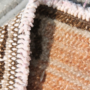 May include: Close-up of a section of a woven rug with a textured surface. The rug features a color palette of brown, beige, and peach. The edge of the rug has a light pink fringe.