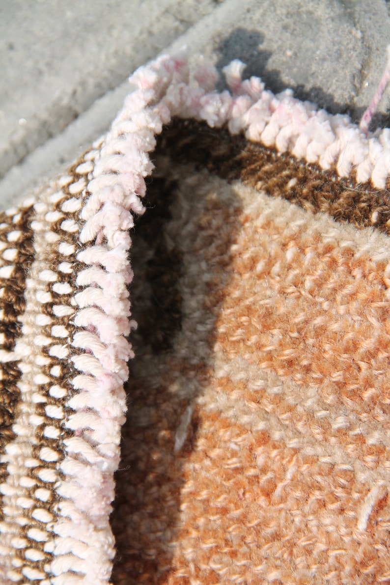 May include: Close-up of a section of a woven rug with a textured surface. The rug features a color palette of brown, beige, and peach. The edge of the rug has a light pink fringe.