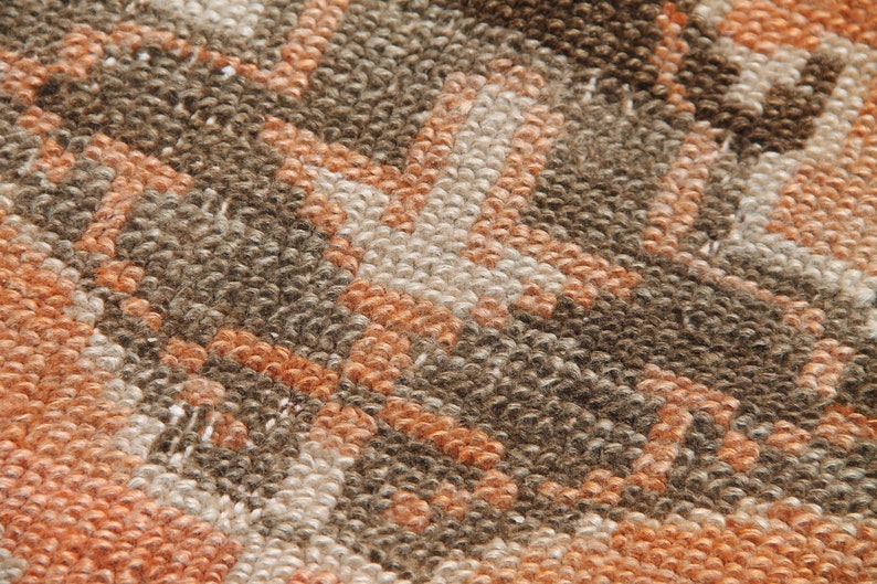 May include: Close-up of a textured rug with a geometric pattern. The rug features a repeating design in shades of brown, orange, and cream. The fibers are tightly woven, creating a dense, durable surface. The rug's design is a blend of modern and traditional styles.