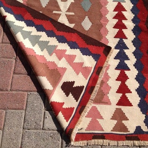 May include: A colorful, geometric patterned rug with a folded edge. The rug features a cream-colored background with red, blue, brown, and pink triangular and diamond shapes. The rug is made of woven fabric and has a fringed edge.
