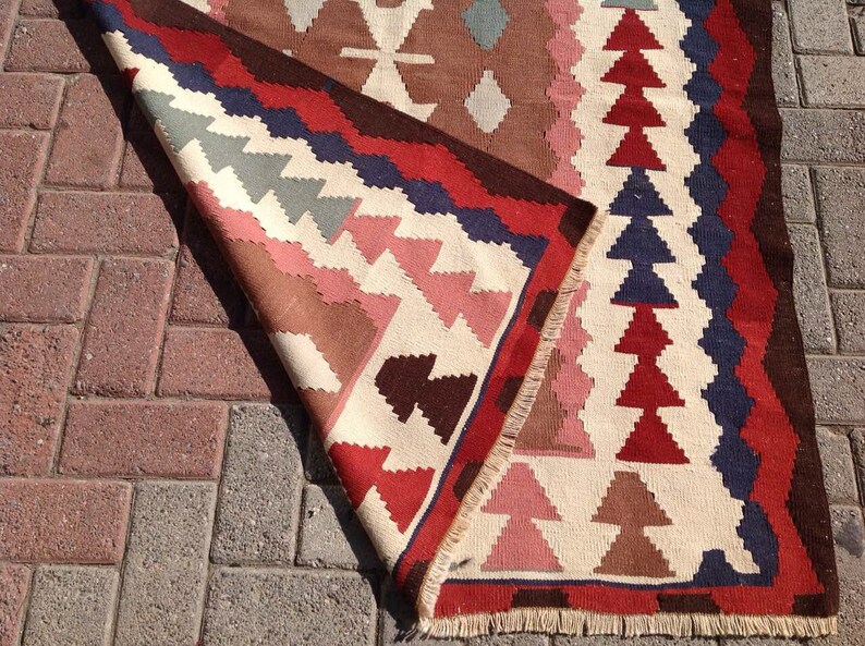 May include: A colorful, geometric patterned rug with a folded edge. The rug features a cream-colored background with red, blue, brown, and pink triangular and diamond shapes. The rug is made of woven fabric and has a fringed edge.