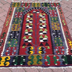 May include: A rectangular, hand-woven rug with a central tree-like design in red, green, and white. The rug features geometric patterns in various colors, including yellow, purple, and black, with a white fringe along the edges.