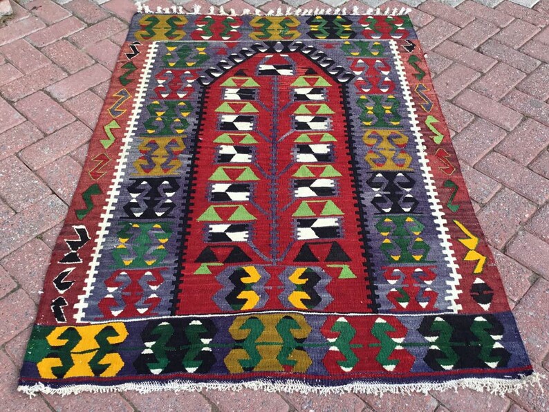 May include: A rectangular, hand-woven rug with a central tree-like design in red, green, and white. The rug features geometric patterns in various colors, including yellow, purple, and black, with a white fringe along the edges.