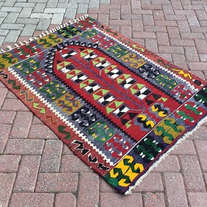 May include: A rectangular, hand-woven rug with a vibrant geometric pattern. The rug features a central red field with a repeating design in white, black, and green. The border includes yellow, green, and black shapes. The rug has a fringed edge.