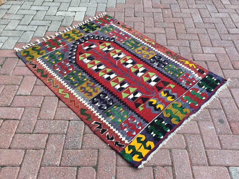 May include: A rectangular, hand-woven rug with a vibrant geometric pattern. The rug features a central red field with a repeating design in white, black, and green. The border includes yellow, green, and black shapes. The rug has a fringed edge.