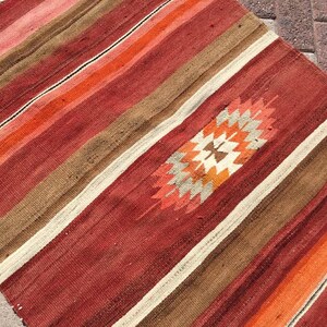 May include: A handwoven rug with a striped pattern in shades of red, orange, brown, and cream. The rug features a central geometric design in orange, cream, and brown. The rug's edges have a fringe.