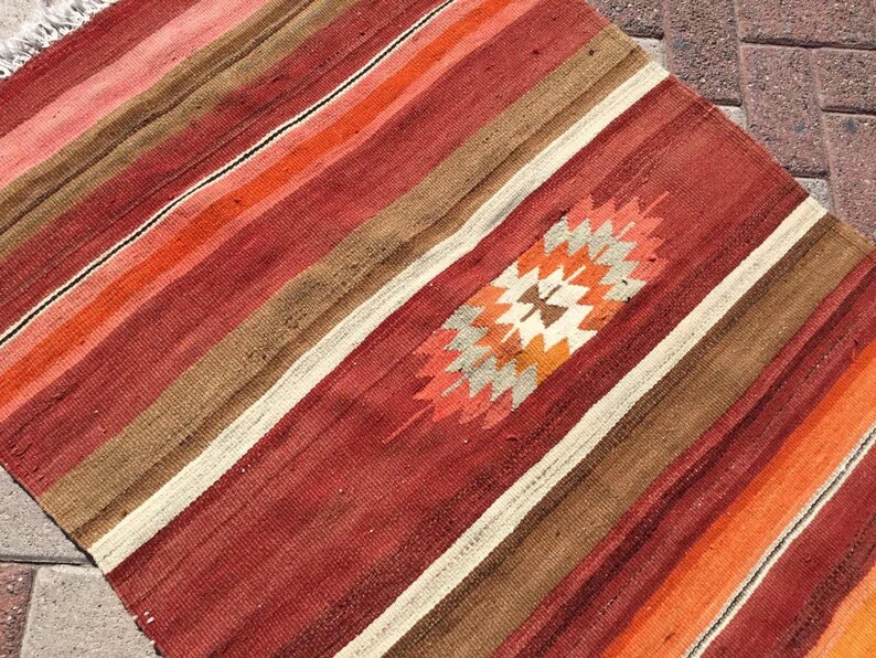 May include: A handwoven rug with a striped pattern in shades of red, orange, brown, and cream. The rug features a central geometric design in orange, cream, and brown. The rug's edges have a fringe.