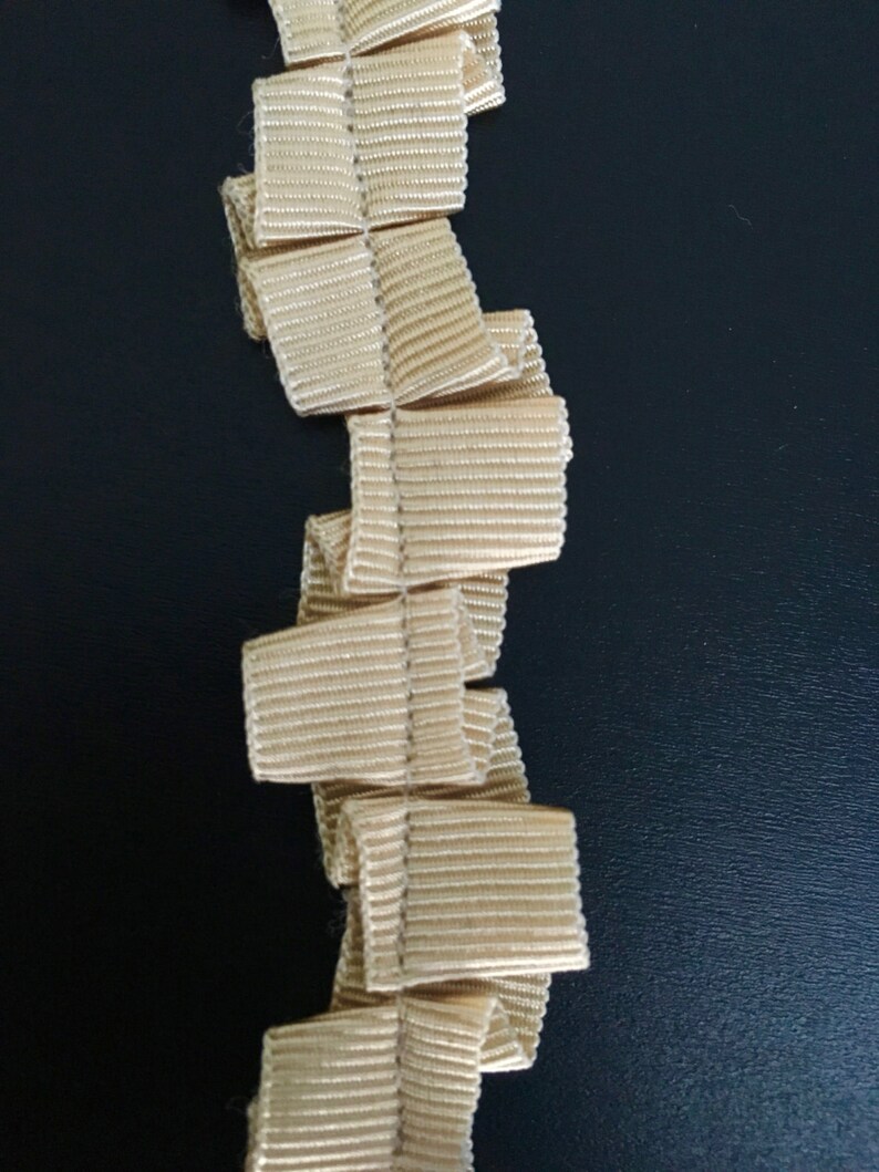 May include: A beige ruffled trim with a textured surface. The trim is folded and stacked, creating a vertical pattern.
