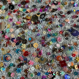 May include: A white mesh fabric adorned with a multitude of colourful sequins and beads. The sequins come in a variety of shapes, including circles, stars, and flowers. The beads are in various colours, including red, blue, green, and purple.