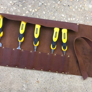 May include: A brown leather tool roll with six slots for chisels. The roll has a strap and is secured with brass rivets. The chisels are yellow and black with the Stanley logo.