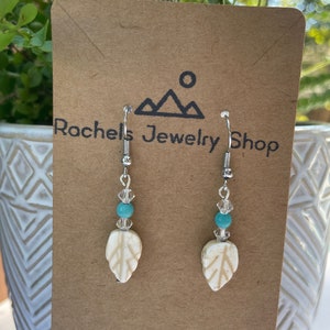 May include: White leaf earrings with turquoise and clear beads on silver hooks. The earrings are displayed on a brown card with the text "Rochel's Jewelry Shop".