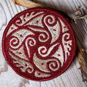 Könnte beinhalten: Runder, gestickter Aufnäher mit einem bordeauxfarbenen Rand und einem cremefarbenen Wirbeldesign. Der Aufnäher hat ein detailliertes, strukturiertes Muster. Ein Stück Holz liegt auf einer hellen Holzoberfläche.