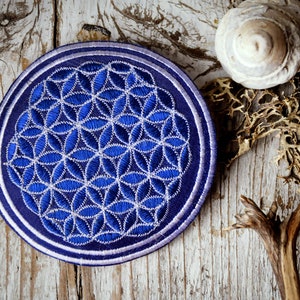 May include: A round, blue embroidered patch featuring a white floral pattern, reminiscent of the Flower of Life symbol. The patch is set against a weathered, light-colored wooden surface, with a seashell and dried plant elements.
