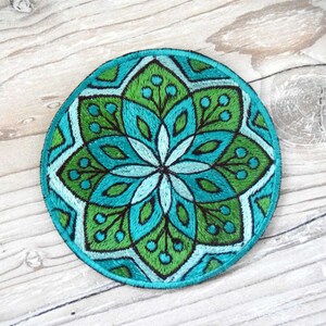 Peut inclure: Écusson rond brodé avec un motif floral symétrique dans les tons de turquoise, vert et blanc. Le patch a une bordure noire et est posé sur un fond de grain de bois clair. Le design est détaillé et visuellement attrayant.