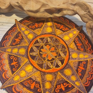 Op de afbeelding: Geborduurde ronde patch met een kompasroosontwerp in bruin, oranje en geel. De patch heeft een gedetailleerd, stervormig patroon met sierlijke versieringen. De patch is waarschijnlijk voor decoratieve doeleinden.