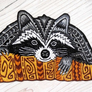 Op de afbeelding: Gestikt patch van een wasbeer met een zwart-wit gezicht, zittend op een gele en bruine gepatroneerde boomstam. De wasbeer heeft een grijs lichaam en een witte neus.