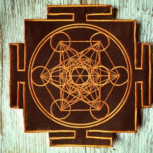 Può includere: Una toppa marrone con un design geometrico dorato ricamato. Il design presenta cerchi, linee e forme interconnesse, con un bordo quadrato e quattro estensioni rettangolari. La toppa è posizionata su una superficie di legno blu chiaro usurata.