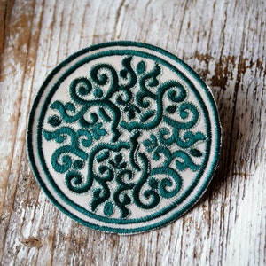 Puede incluir: Un parche redondo de tela blanca con un diseño bordado en verde de un patrón floral estilizado.