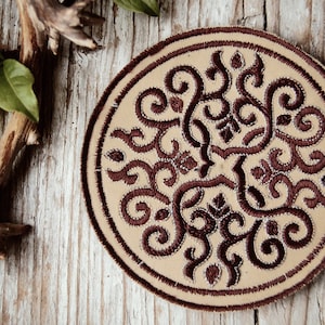 Puede incluir: Un parche redondo de tela beige con un diseño bordado marrón de espirales y patrones intrincados. El parche está sobre una superficie de madera.
