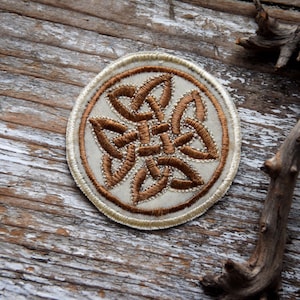 Könnte beinhalten: Ein runder, weißer Stoff-Patch mit einem braunen, gestickten keltischen Knotendesign.