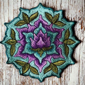 Pode incluir: Um remendo bordado de flores turquesa e roxo com folhas verdes. O remendo é circular e tem um design tipo mandala.